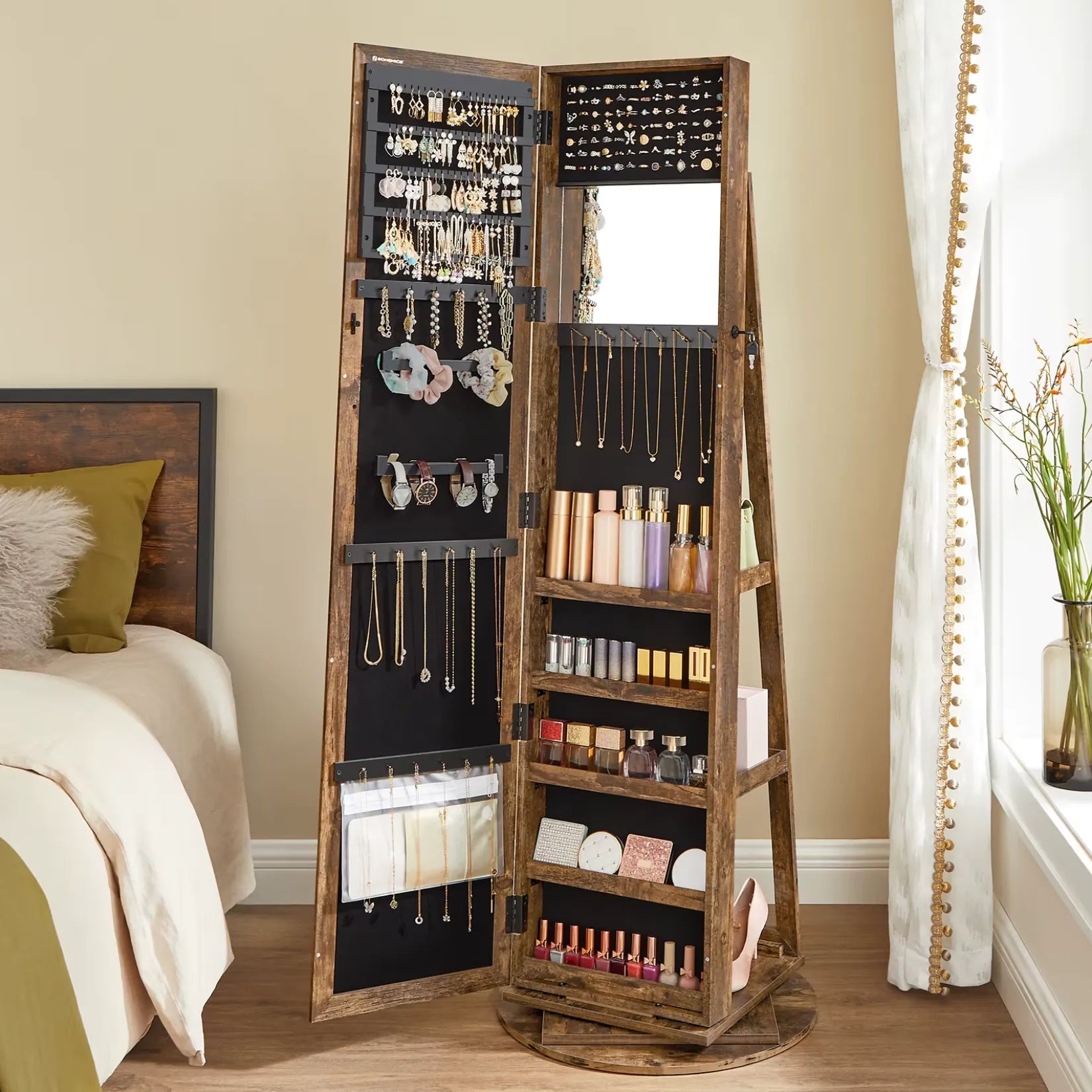 Armoire à bijoux sur pied bois marron ouverte dans une chambre lumineuse avec rideaux blancs.