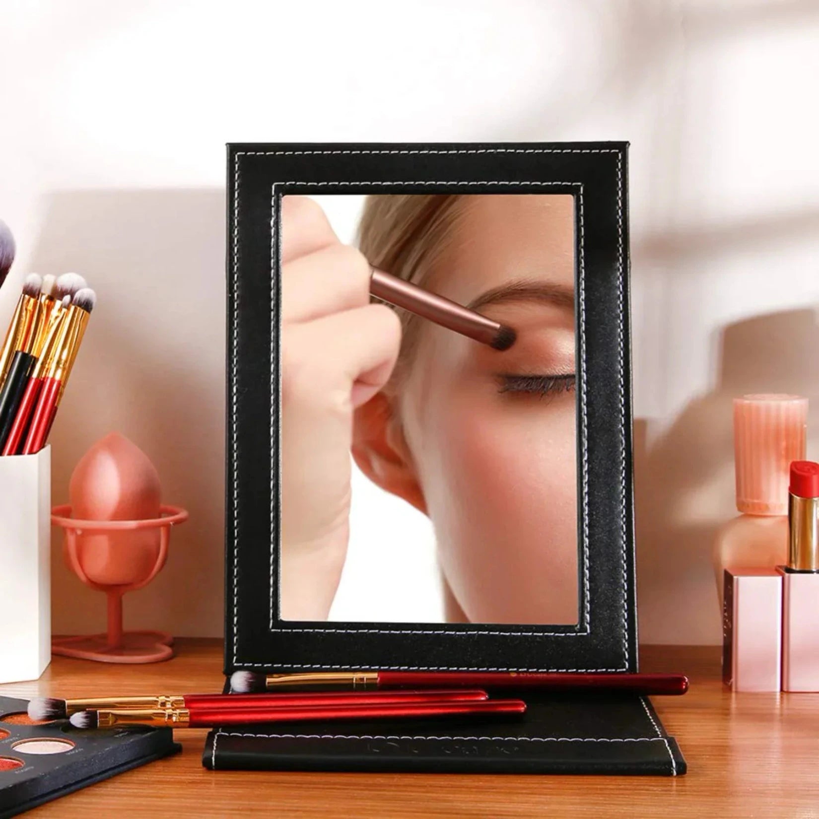 Miroir de poche cuir noir utilisé sur une table de maquillage avec pinceaux.
