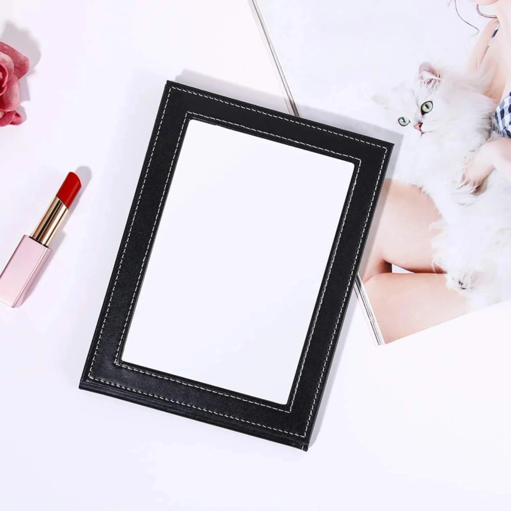 Miroir de poche cuir noir posé à plat à côté d’un rouge à lèvres et d’un magazine.