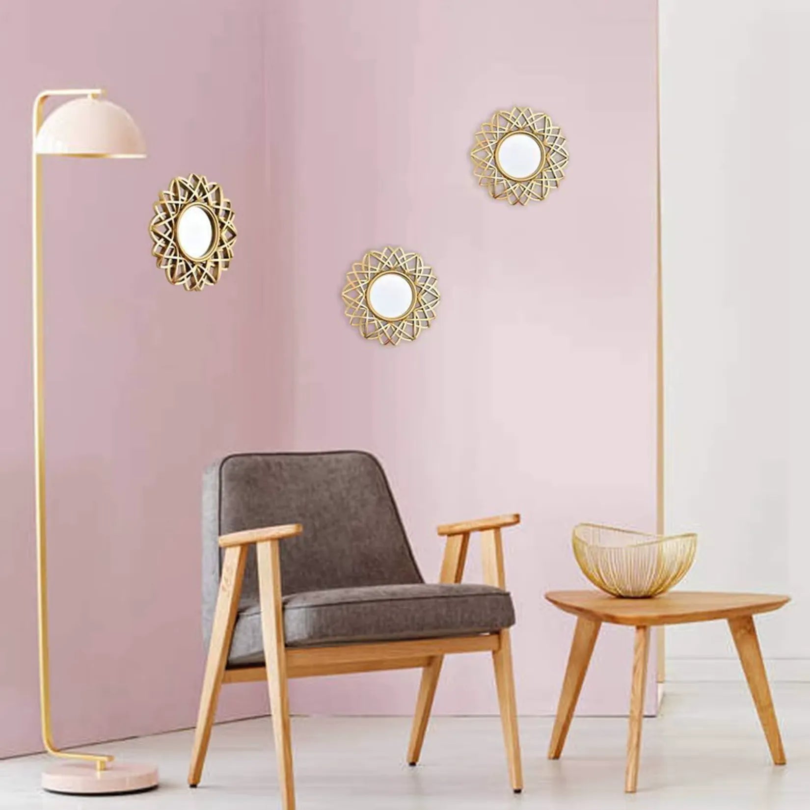 Miroir rond design doré accroché dans un salon scandinave aux murs roses avec fauteuil gris clair et lampe vintage.