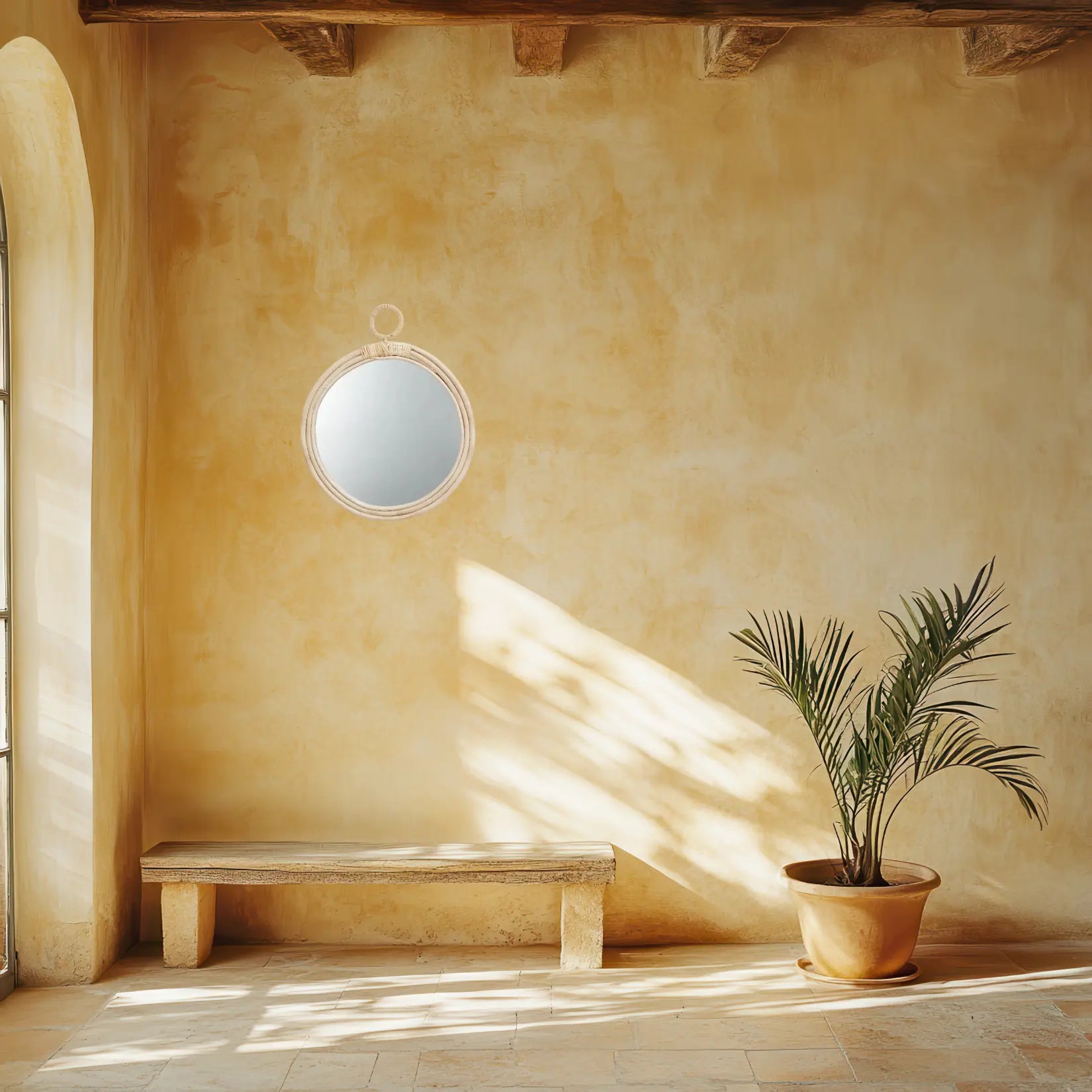 Miroir Rotin Rond avec anneau accroché sur un mur ocre lumineux, ambiance méditerranéenne avec banc en pierre et plante verte.